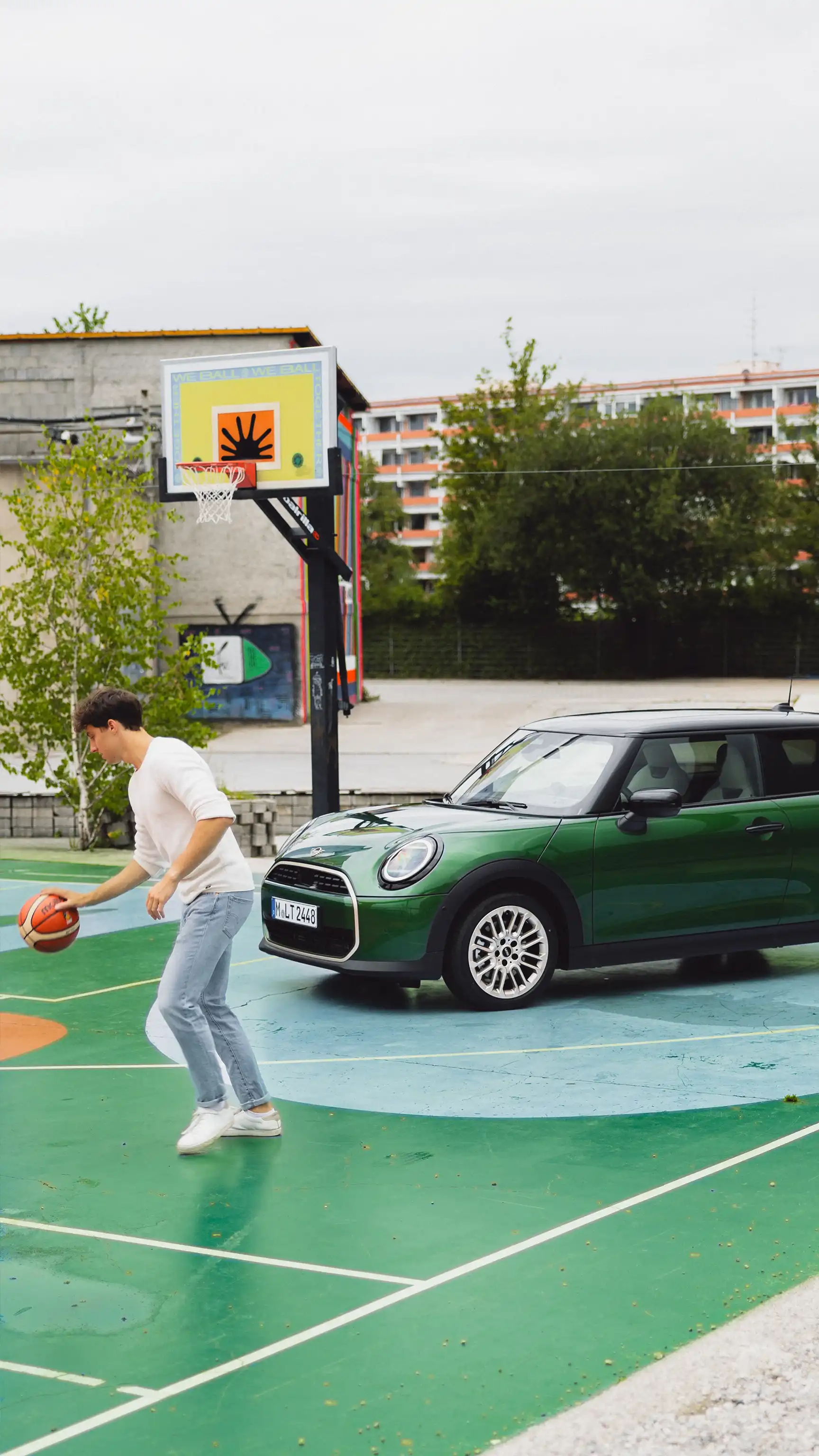 Person dribbling a basketball on an outdoor court next to a MINI Cooper in Racing Green IV.
