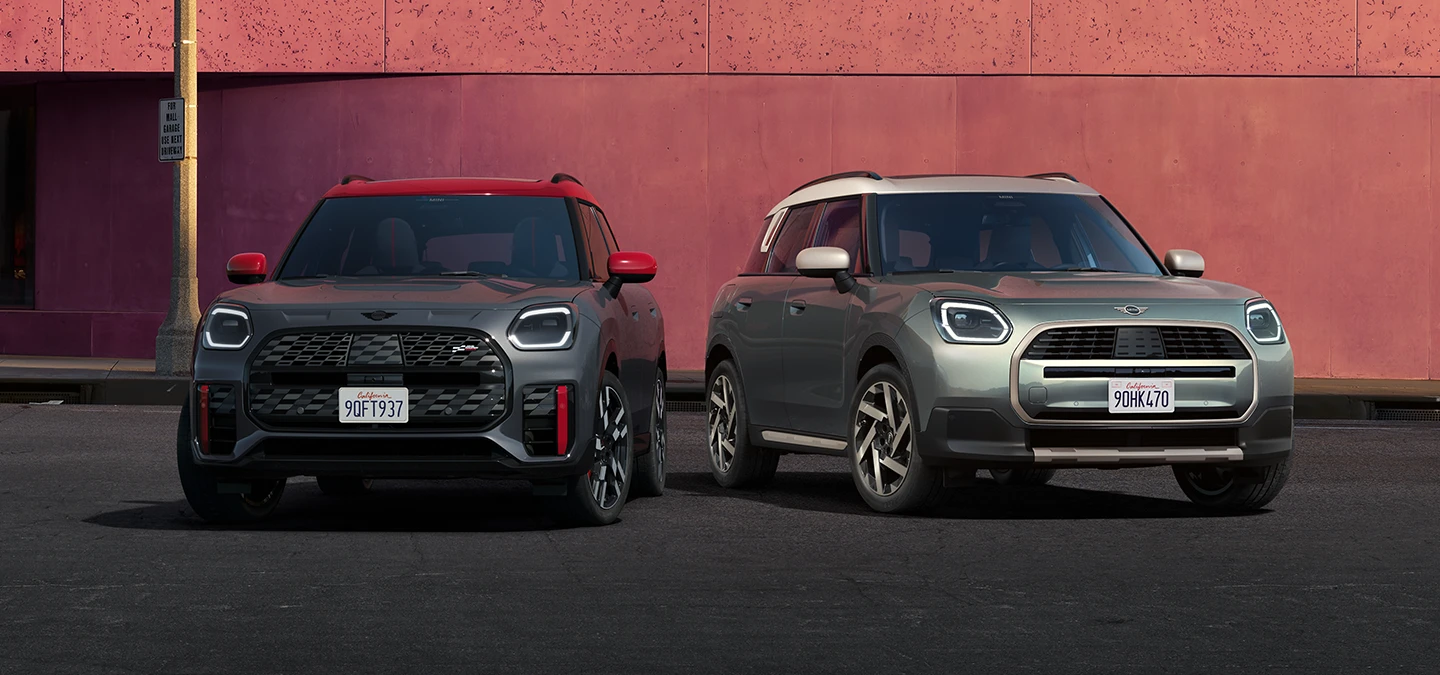 Front view of the MINI Countryman JCW in Legend Grey and Red Roof and the MINI Countryman in Smokey Green and Vibrant Silver Roof standing next to each other.